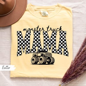 Op de afbeelding: Boterkleurig T-shirt met de tekst "monster truck MAMA" in zwart, met een geblokt patroon in de letters. Een zwarte monster truck afbeelding staat onder de tekst. Een bruine hoed en droogbloemen staan op de achtergrond.