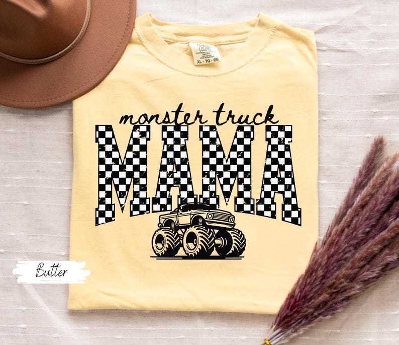 Op de afbeelding: Boterkleurig T-shirt met de tekst "monster truck MAMA" in zwart, met een geblokt patroon in de letters. Een zwarte monster truck afbeelding staat onder de tekst. Een bruine hoed en droogbloemen staan op de achtergrond.