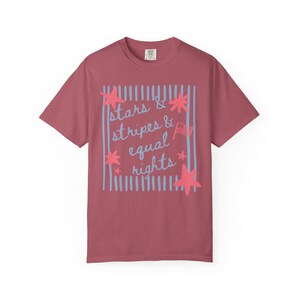 Puede incluir: Camiseta granate con las palabras "stars & stripes & equal rights" en escritura azul claro y un borde a rayas azul claro a juego. Estrellas rosas y una peque&ntilde;a bandera americana.