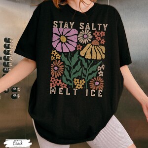 Op de afbeelding: Zwart T-shirt met een bloemenontwerp en de tekst "STAY SALTY MELT ICE". De afbeelding toont verschillende bloemen in roze, geel en oranje met groene bladeren. Het shirt heeft een casual, oversized pasvorm.