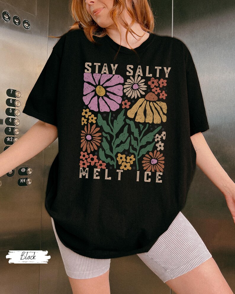 Op de afbeelding: Zwart T-shirt met een bloemenontwerp en de tekst "STAY SALTY MELT ICE". De afbeelding toont verschillende bloemen in roze, geel en oranje met groene bladeren. Het shirt heeft een casual, oversized pasvorm.