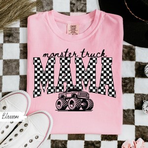 May include: Pink Comfort Colors t-shirt with the words "monster truck MAMA" in a black and white checkered design. A black monster truck graphic is centered below the text. White sneakers are in the lower left corner.