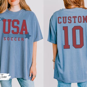 May include: A light blue t-shirt with the text "USA SOCCER" in red. The front of the shirt has a star graphic. The back of the shirt has the word "CUSTOM" above the number "10" in red.