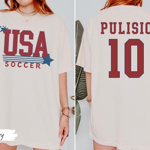 May include: Ivory-colored t-shirt with "USA SOCCER" in red and blue text, featuring stars and stripes design. The back displays "PULISIC 10" in a similar color scheme. A casual, patriotic soccer-themed apparel.