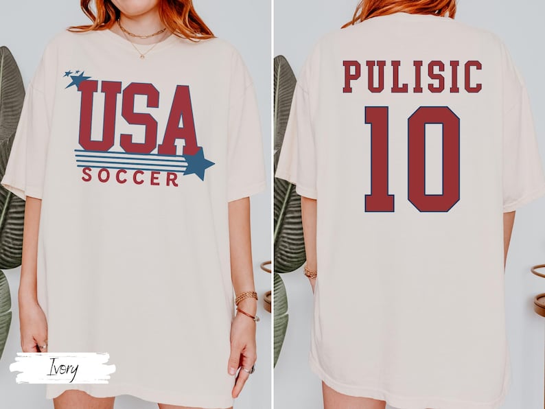 May include: Ivory-colored t-shirt with "USA SOCCER" in red and blue text, featuring stars and stripes design. The back displays "PULISIC 10" in a similar color scheme. A casual, patriotic soccer-themed apparel.