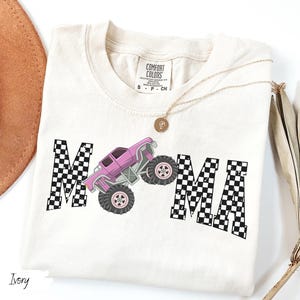 May include: Ivory-colored t-shirt featuring the word "MAMA" with a checkered pattern and a pink monster truck graphic. The shirt has a crew neck and short sleeves. A gold necklace with a pendant is draped on the shirt.