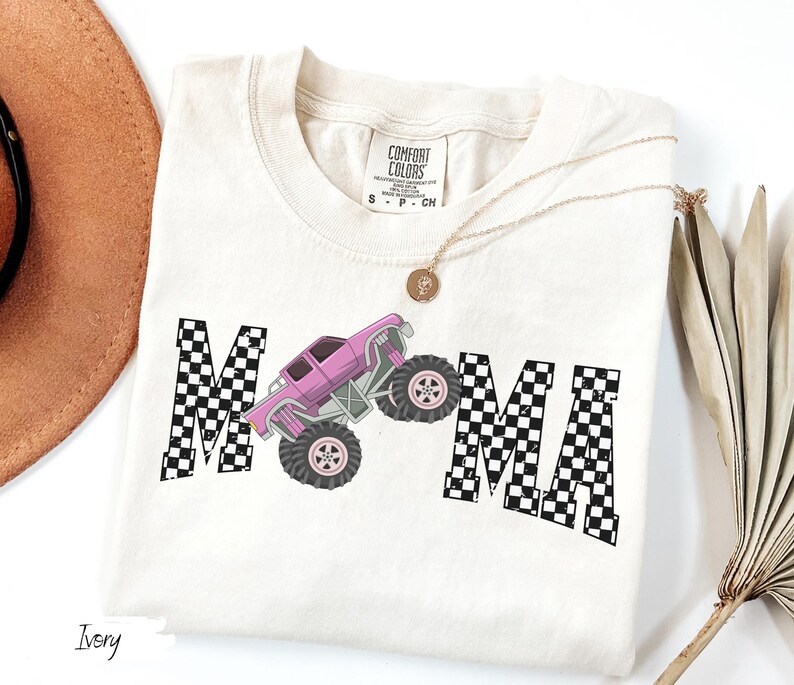May include: Ivory-colored t-shirt featuring the word "MAMA" with a checkered pattern and a pink monster truck graphic. The shirt has a crew neck and short sleeves. A gold necklace with a pendant is draped on the shirt.