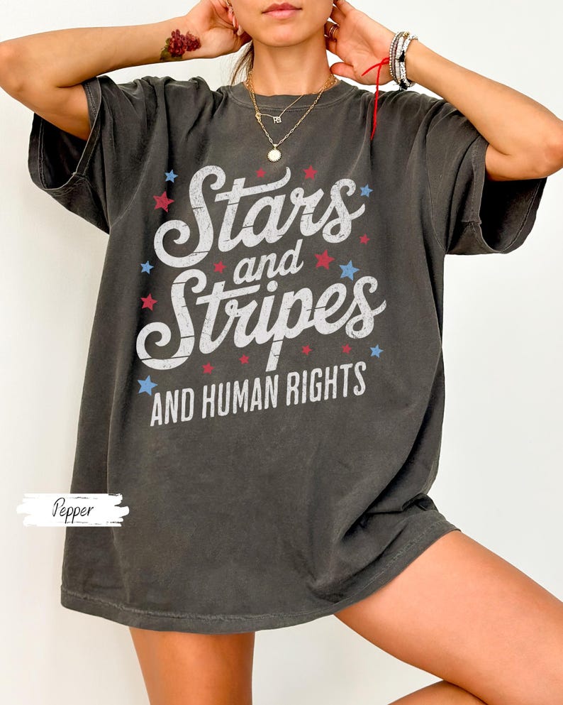 Puede incluir: Camiseta gris oscuro con las palabras "Stars and Stripes AND HUMAN RIGHTS" en escritura blanca y estrellas rojas y azules. La camiseta es de corte holgado.