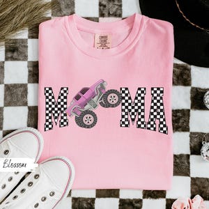 May include: A light pink t-shirt with the word "MAMA" in a checkered pattern. A purple monster truck graphic is between the letters. White sneakers are in the bottom left corner.