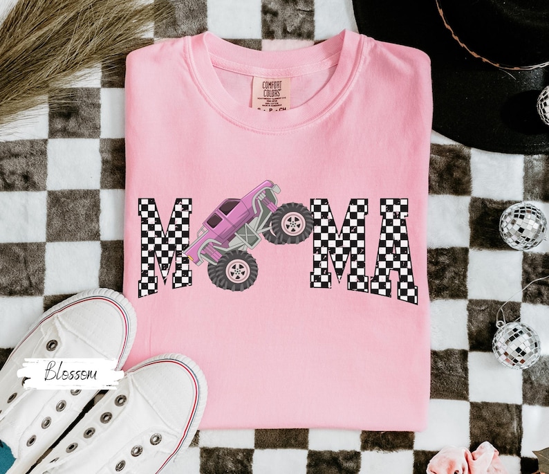 May include: A light pink t-shirt with the word "MAMA" in a checkered pattern. A purple monster truck graphic is between the letters. White sneakers are in the bottom left corner.