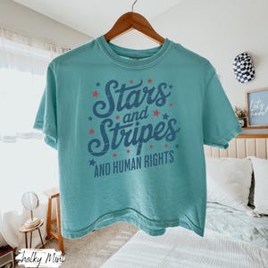 Puede incluir: Una camiseta turquesa con la frase "Stars and Stripes AND HUMAN RIGHTS" en azul marino, adornada con estrellas rojas. La camiseta est&aacute; colgada de una percha de madera. El dise&ntilde;o tiene un estilo vintage.
