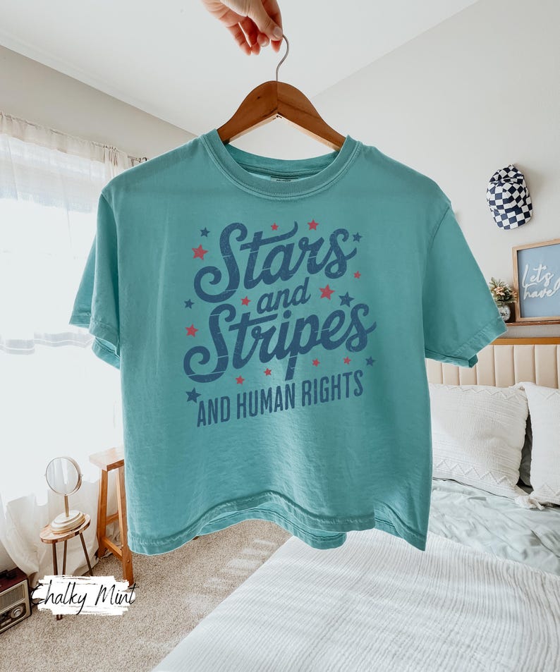 Puede incluir: Una camiseta turquesa con la frase "Stars and Stripes AND HUMAN RIGHTS" en azul marino, adornada con estrellas rojas. La camiseta est&aacute; colgada de una percha de madera. El dise&ntilde;o tiene un estilo vintage.