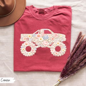 Op de afbeelding: Karmijnrood T-shirt met een bloemen monster truck ontwerp. De carrosserie en wielen van de truck zijn gevuld met roze, witte en blauwe bloemen. Het shirt is opgevouwen en gepresenteerd op een wit oppervlak. Het woord "Crimson" staat linksonder.