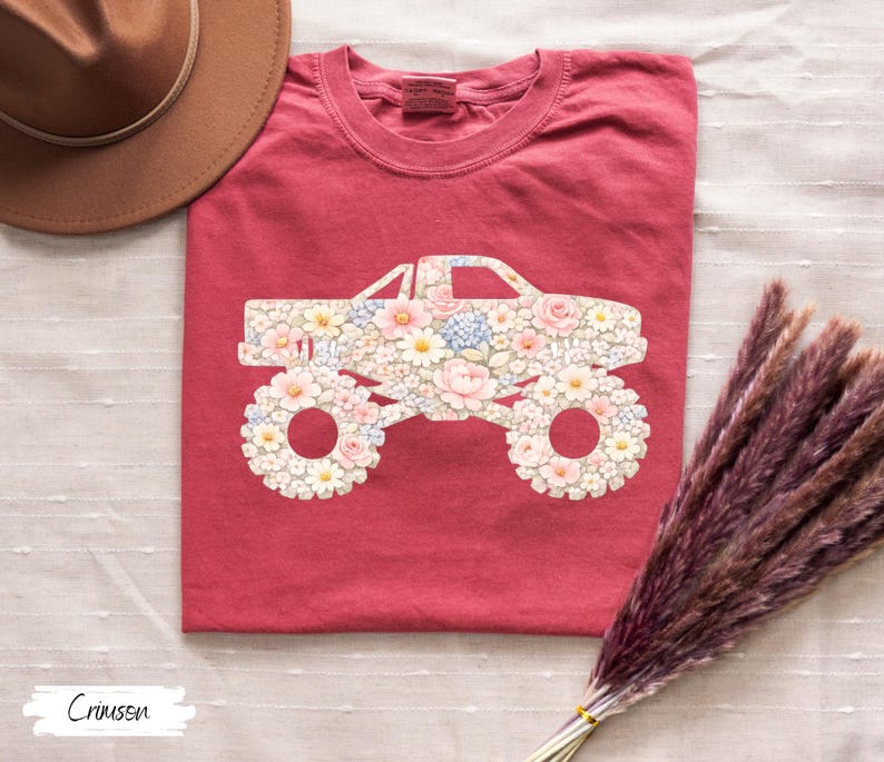 Op de afbeelding: Karmijnrood T-shirt met een bloemen monster truck ontwerp. De carrosserie en wielen van de truck zijn gevuld met roze, witte en blauwe bloemen. Het shirt is opgevouwen en gepresenteerd op een wit oppervlak. Het woord "Crimson" staat linksonder.