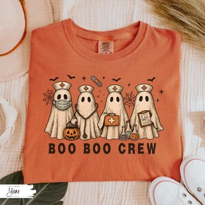 May include: An orange t-shirt featuring a graphic of four ghost characters dressed as medical professionals. The ghosts wear nurse hats, and hold medical tools. The text "BOO BOO CREW" is printed in black block letters below the ghosts.