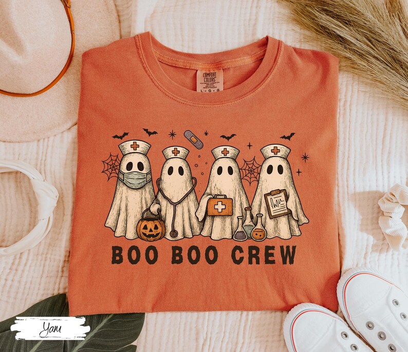 May include: An orange t-shirt featuring a graphic of four ghost characters dressed as medical professionals. The ghosts wear nurse hats, and hold medical tools. The text "BOO BOO CREW" is printed in black block letters below the ghosts.
