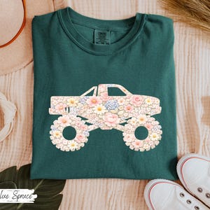 Op de afbeelding: Een donkergroen t-shirt met een bloemenmonster truck ontwerp. De truck is gevuld met roze, witte en gele bloemen. De tekst "Blue Spruce" staat linksonder.