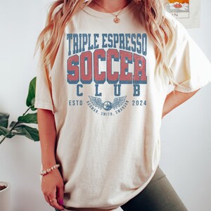 Op de afbeelding: Een vrouw draagt een lichtbeige T-shirt met de tekst "Triple Espresso Soccer Club" in rood en blauw. Het shirt heeft ook een blauw en wit logo met vleugels en een voetbal. De tekst "Estd 2024" staat onder het logo. De vrouw draagt donkergroene fietsbroeken.