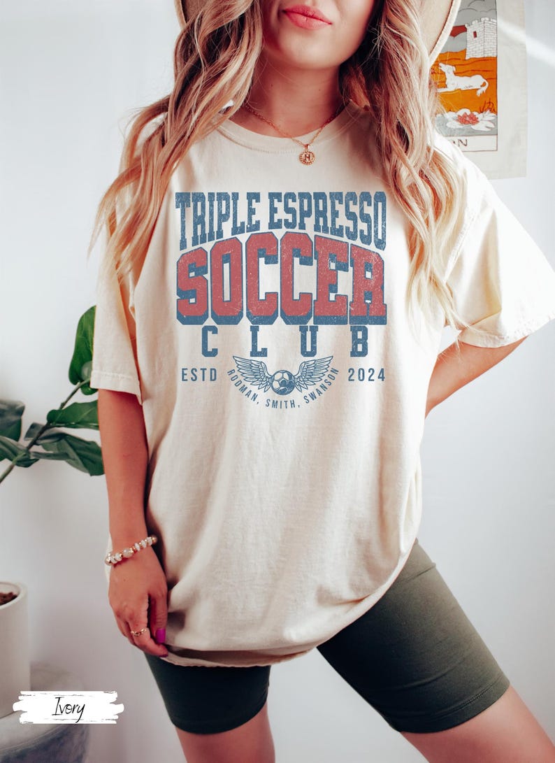 Op de afbeelding: Een vrouw draagt een lichtbeige T-shirt met de tekst "Triple Espresso Soccer Club" in rood en blauw. Het shirt heeft ook een blauw en wit logo met vleugels en een voetbal. De tekst "Estd 2024" staat onder het logo. De vrouw draagt donkergroene fietsbroeken.
