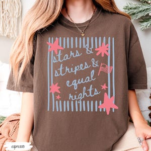 Puede incluir: Camiseta color espresso con el texto "stars & stripes & equal rights" en un dise&ntilde;o azul y rosa. La camiseta presenta un marco rectangular con rayas azules verticales y acentos de estrellas rosas.