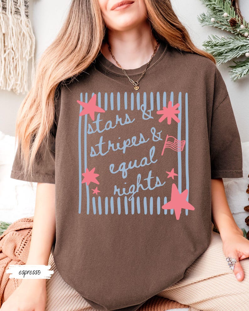 Puede incluir: Camiseta color espresso con el texto "stars & stripes & equal rights" en un dise&ntilde;o azul y rosa. La camiseta presenta un marco rectangular con rayas azules verticales y acentos de estrellas rosas.
