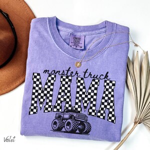 Op de afbeelding: Een violet Comfort Colors t-shirt met de tekst "monster truck MAMA" in zwart, met een geblokt patroon in de letters. Een afbeelding van een monstertruck staat eronder. Een gouden ketting en een bruine hoed staan ook in de afbeelding.