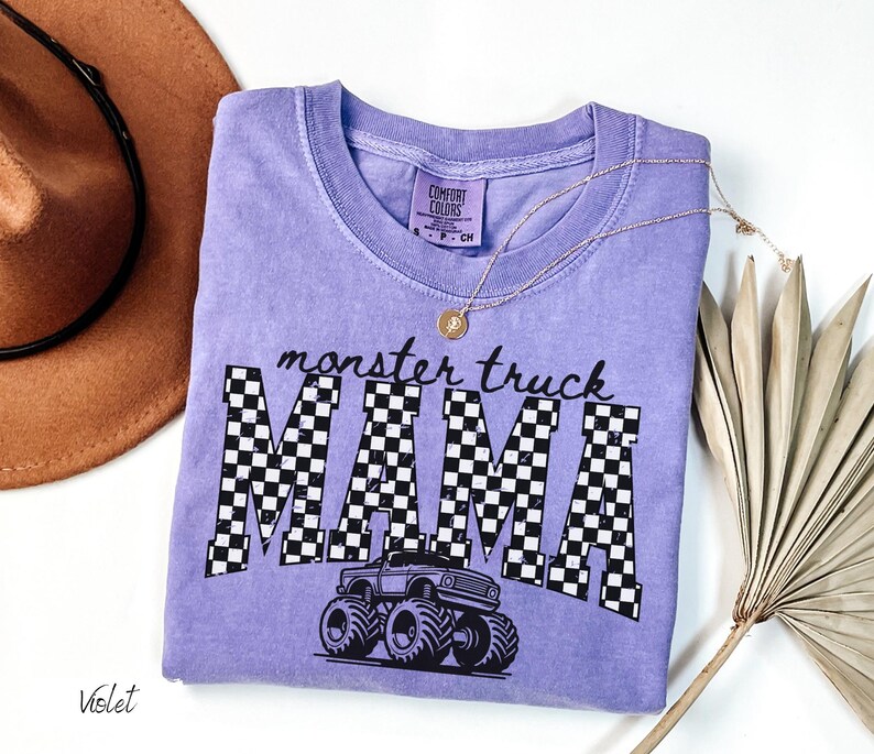 Op de afbeelding: Een violet Comfort Colors t-shirt met de tekst "monster truck MAMA" in zwart, met een geblokt patroon in de letters. Een afbeelding van een monstertruck staat eronder. Een gouden ketting en een bruine hoed staan ook in de afbeelding.