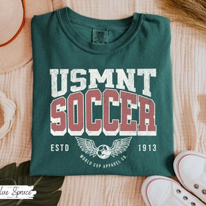 Może przedstawiać: Ciemnozielona koszulka z napisem "USMNT SOCCER" w białych i czerwonych literach. Poniżej piłka nożna ze skrzydłami i słowami "ESTD 1913". Koszulka jest złożona na jasnej powierzchni.