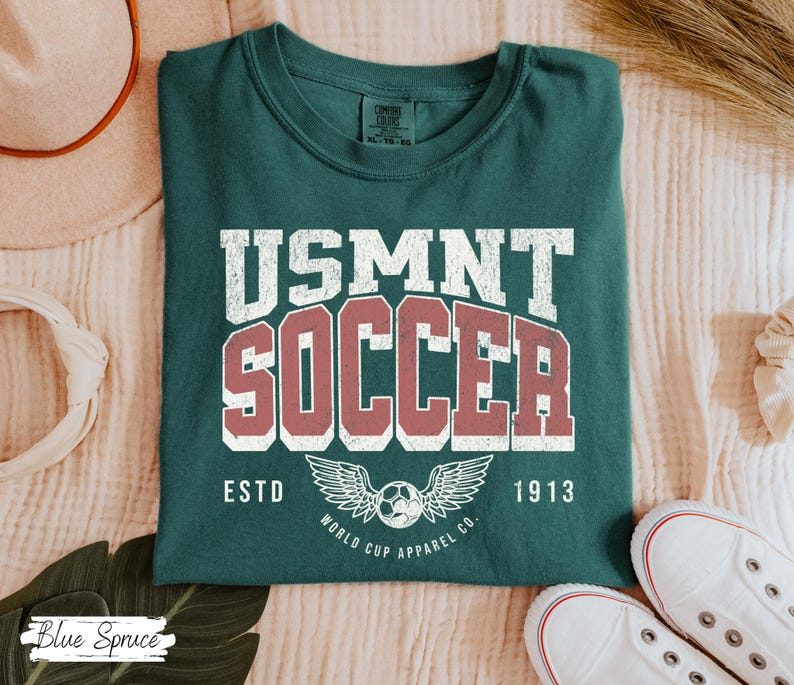 Może przedstawiać: Ciemnozielona koszulka z napisem "USMNT SOCCER" w białych i czerwonych literach. Poniżej piłka nożna ze skrzydłami i słowami "ESTD 1913". Koszulka jest złożona na jasnej powierzchni.