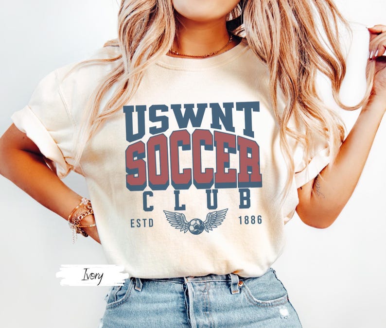 May include: A white t-shirt with the text "USWNT SOCCER CLUB ESTD 1886" in blue and red lettering. The shirt features a blue and white winged soccer ball graphic.