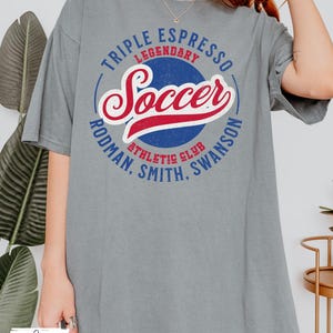 Puede incluir: Una camiseta gris con un dise&ntilde;o gr&aacute;fico de estilo vintage con las palabras "Triple Espresso Legendary Soccer Rodman, Smith, Swanson Athletic Club".