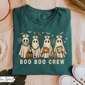 May include: Teal-colored t-shirt featuring a graphic of four ghost characters dressed as medical professionals. The ghosts are wearing medical hats, masks, and carrying medical tools. The text "BOO BOO CREW" is printed below the ghosts.