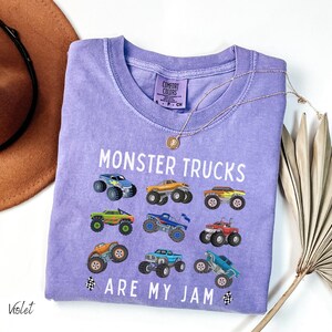 May include: A violet Comfort Colors t-shirt featuring the text "MONSTER TRUCKS ARE MY JAM" and illustrations of monster trucks. The shirt has a crew neck and short sleeves. A gold necklace is draped over the shirt.
