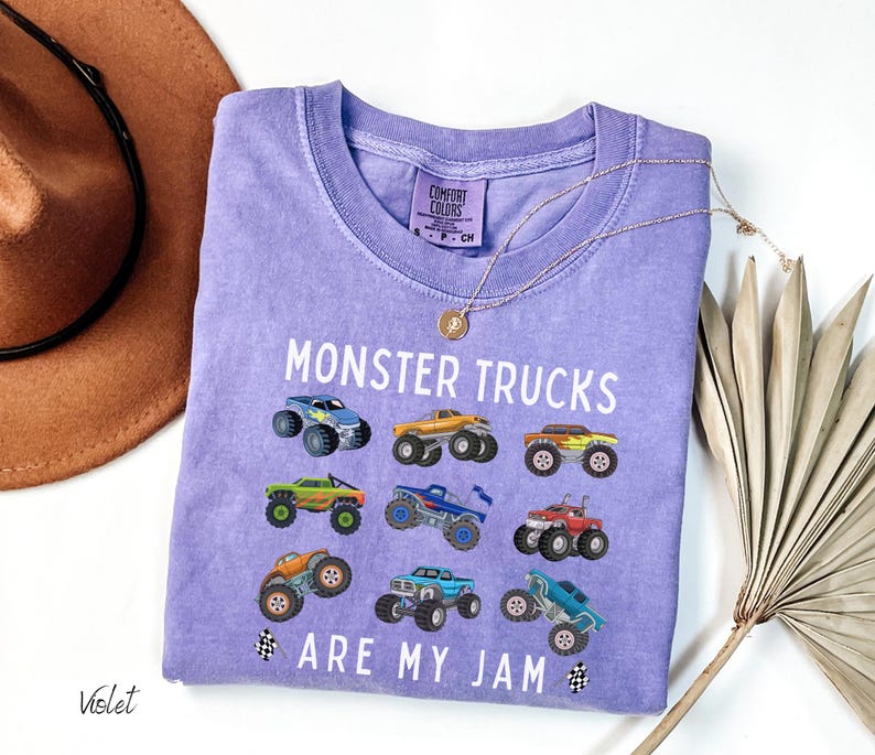 May include: A violet Comfort Colors t-shirt featuring the text "MONSTER TRUCKS ARE MY JAM" and illustrations of monster trucks. The shirt has a crew neck and short sleeves. A gold necklace is draped over the shirt.