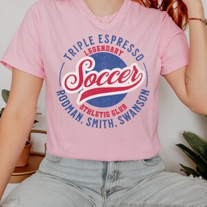 Puede incluir: Una camiseta rosa con un gr&aacute;fico azul y rojo que dice "Triple Espresso Legendary Soccer Rodman, Smith, Swanson Athletic Club".