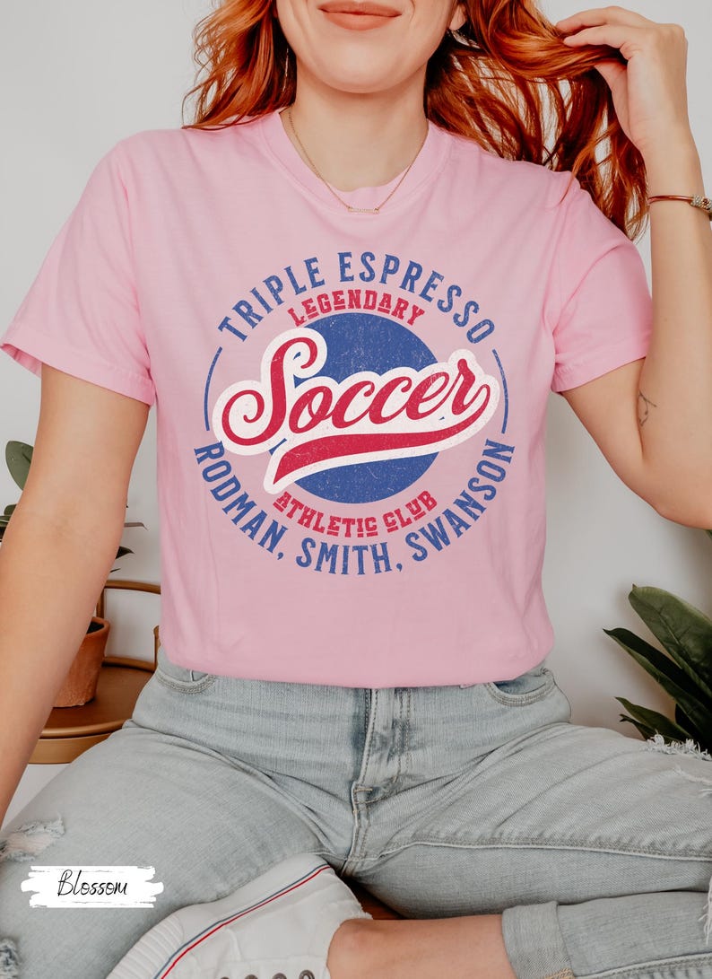 Puede incluir: Una camiseta rosa con un gr&aacute;fico azul y rojo que dice "Triple Espresso Legendary Soccer Rodman, Smith, Swanson Athletic Club".
