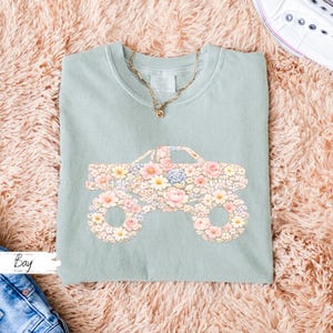 Op de afbeelding: Een lichtgroen t-shirt met een bloemenmonster truck ontwerp. De truck is gevuld met roze, gele en blauwe bloemen. Een gouden ketting is zichtbaar bij de halslijn. Het woord "Bay" staat linksonder.