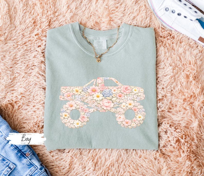 Op de afbeelding: Een lichtgroen t-shirt met een bloemenmonster truck ontwerp. De truck is gevuld met roze, gele en blauwe bloemen. Een gouden ketting is zichtbaar bij de halslijn. Het woord "Bay" staat linksonder.
