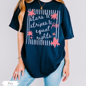 Puede incluir: Camiseta azul marino con el texto "Stars & Stripes & Equal Rights" en un dise&ntilde;o blanco y rosa. El dise&ntilde;o incluye estrellas, rayas y una peque&ntilde;a bandera americana. La camiseta es de corte holgado.