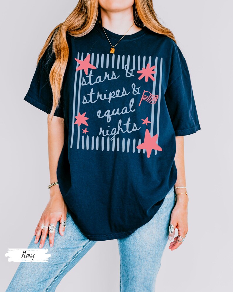 Puede incluir: Camiseta azul marino con el texto "Stars & Stripes & Equal Rights" en un dise&ntilde;o blanco y rosa. El dise&ntilde;o incluye estrellas, rayas y una peque&ntilde;a bandera americana. La camiseta es de corte holgado.