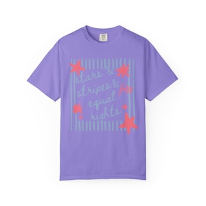 Puede incluir: Una camiseta de color lila con la frase "stars & stripes & equal rights" en una fuente manuscrita. El dise&ntilde;o incluye estrellas rojas y rayas azul claro, creando un tema patri&oacute;tico.