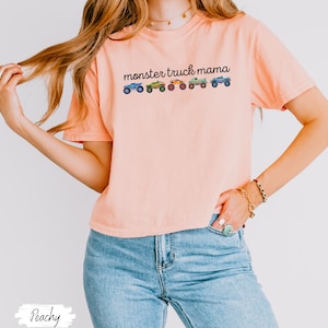 May include: Peach-colored t-shirt with the text "monster truck mama" above a row of colorful monster truck illustrations. The shirt is made of a soft material and has short sleeves.