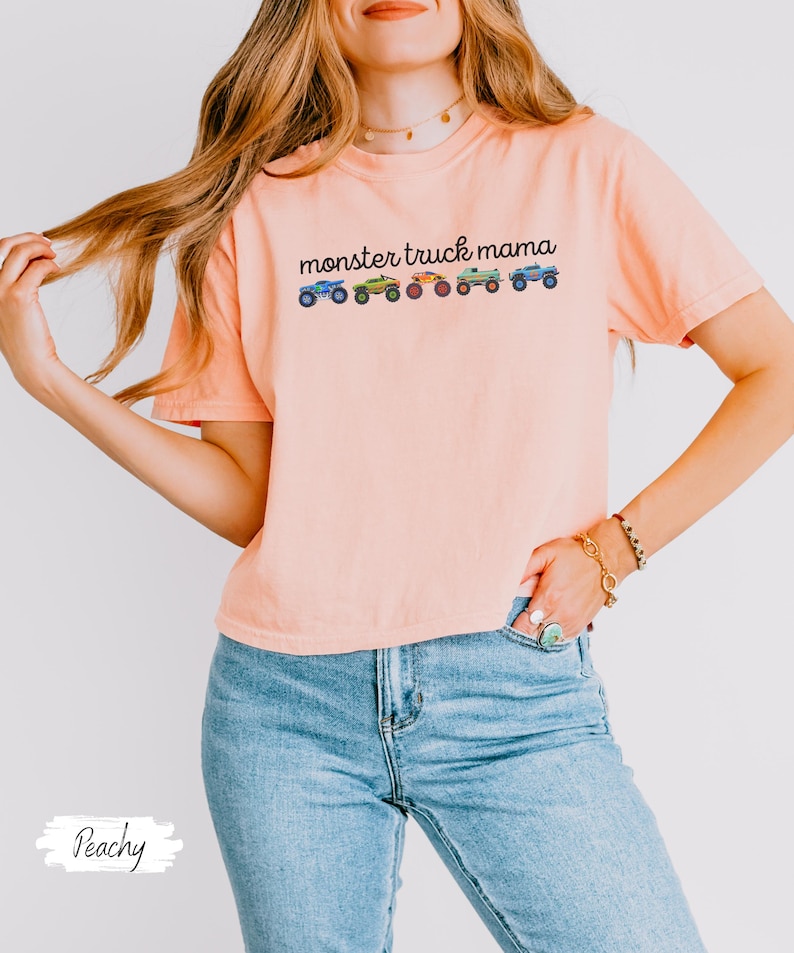 May include: Peach-colored t-shirt with the text "monster truck mama" above a row of colorful monster truck illustrations. The shirt is made of a soft material and has short sleeves.
