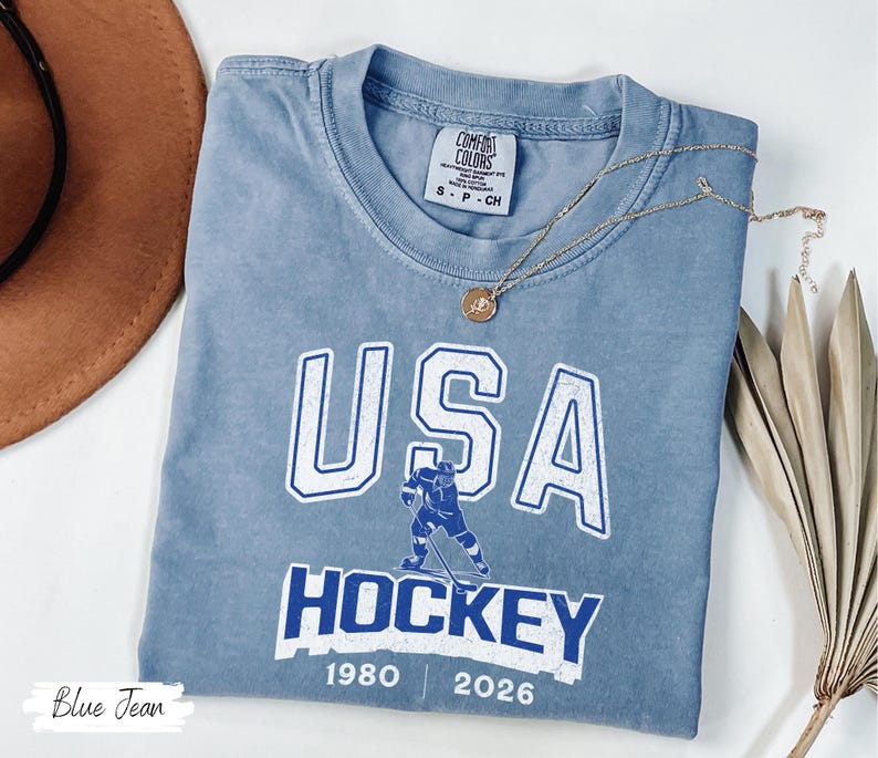 May include: A blue Comfort Colors t-shirt with a USA Hockey graphic. The design features the word "USA" in white block letters, a hockey player illustration, and the word "HOCKEY" in white. The shirt also includes the years 1980 and 2026.