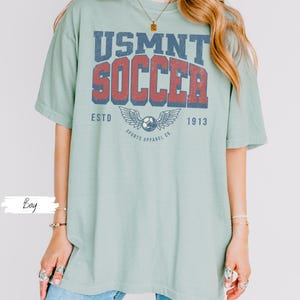 May include: A pale green t-shirt featuring the text "USMNT SOCCER" in blue and red, with a soccer ball and wing design. The shirt also includes the text "ESTD 1913 SPORTS APPAREL CO."