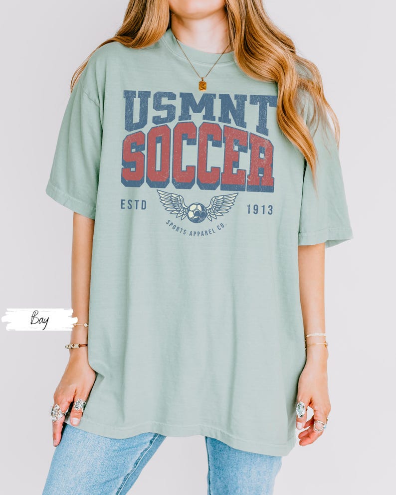 May include: A pale green t-shirt featuring the text "USMNT SOCCER" in blue and red, with a soccer ball and wing design. The shirt also includes the text "ESTD 1913 SPORTS APPAREL CO."