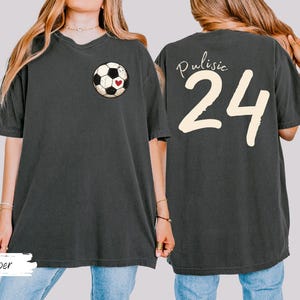 Op de afbeelding: Donkergrijs T-shirt met een voetbalafbeelding en een hart op de voorkant. De achterkant van het shirt toont de naam "Pulisic" boven het nummer "24" in cr&egrave;me. Het woord "Pepper" staat onderaan.