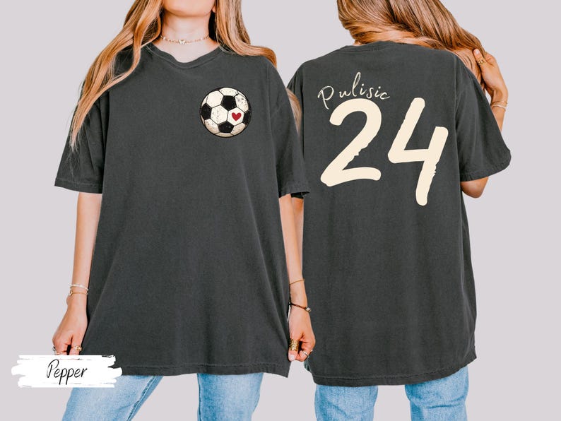 Op de afbeelding: Donkergrijs T-shirt met een voetbalafbeelding en een hart op de voorkant. De achterkant van het shirt toont de naam "Pulisic" boven het nummer "24" in cr&egrave;me. Het woord "Pepper" staat onderaan.