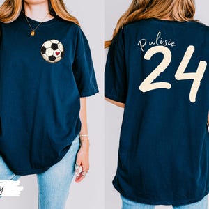 Op de afbeelding: Marineblauw T-shirt met een voetbalafbeelding en een rood hart op de voorkant. De achterkant van het shirt toont de naam "Pulisic" boven het nummer "24" in cr&egrave;me. Een casual, comfortabele stijl.