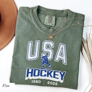 May include: An olive green t-shirt with the words "USA HOCKEY" in white and blue lettering. A hockey player graphic is featured below the word "USA". The shirt also has the years "1980" and "2026" printed on it. A gold necklace is draped across the shirt.